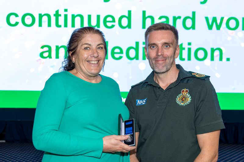 Two East Midlands Ambulance Service colleagues stand together on stage during a long service awards presentation. One colleague holds an open medal presentation case while both pose for a commemorative photograph in front of a large display screen.