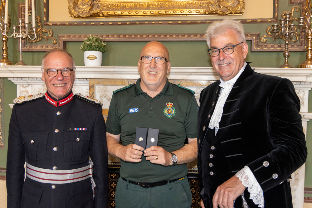 Andrew Britton stands in between the Lord-Lieutenant of Northamptonshire and High Sheriff of Northamptonshire. He is in green ambulance uniform and holds up his award.