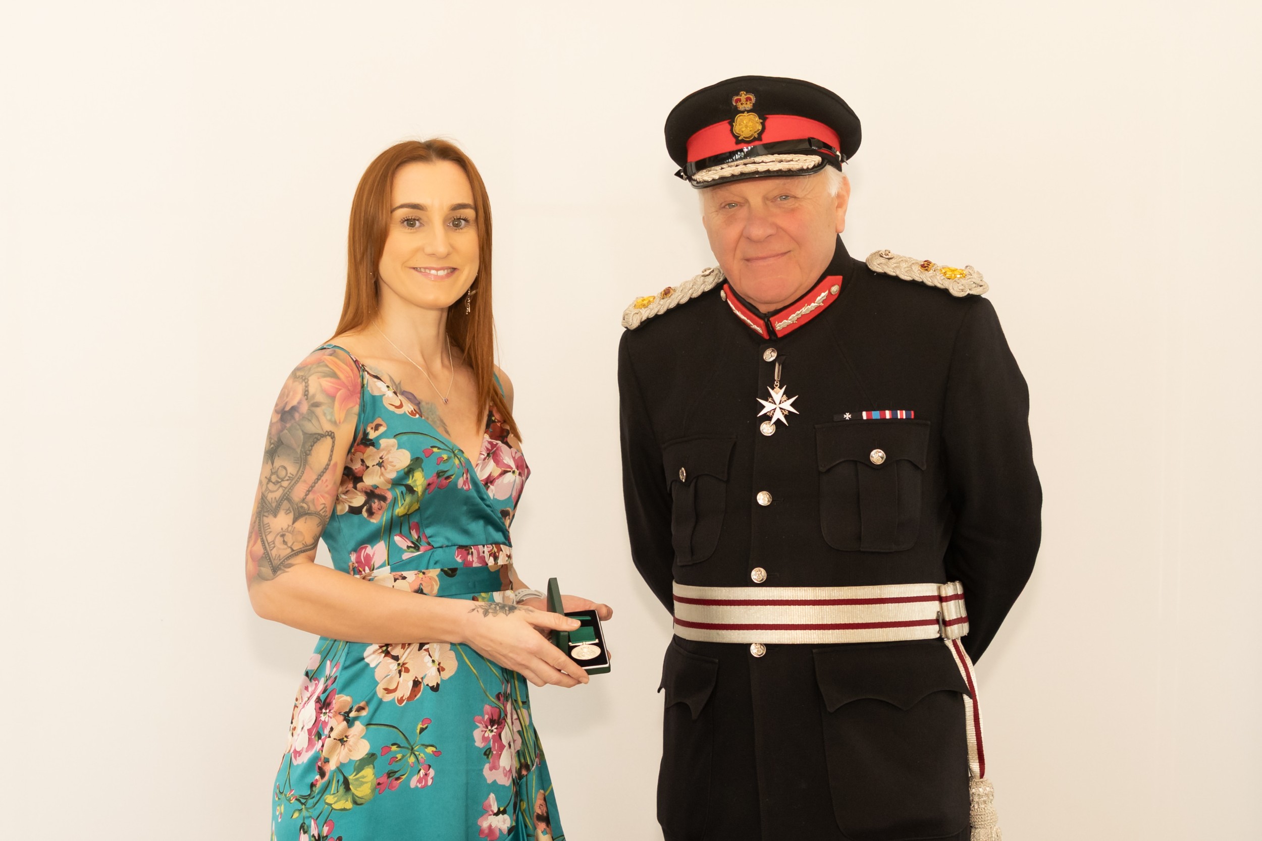 Danielle Hitchen in a floral dress stands beside the Lord-Lieutenant Mr Toby Dennis, who is wearing formal ceremonial uniform with medals and a peaked cap. Danielle his holding a small presentation box, and both are standing against a plain light-coloured background.