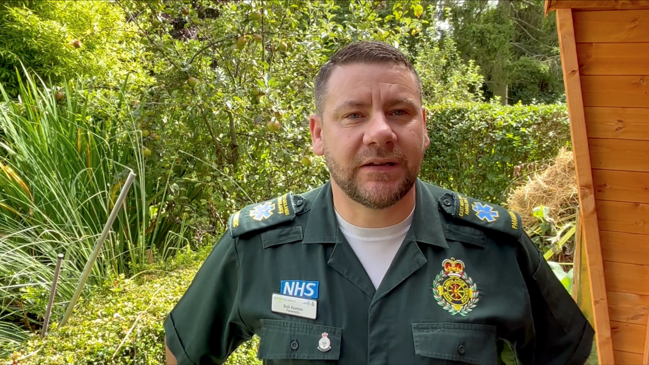 Rob Keenan stood in a garden surrounded by green plants. He is wearing a green ambulance shirt.