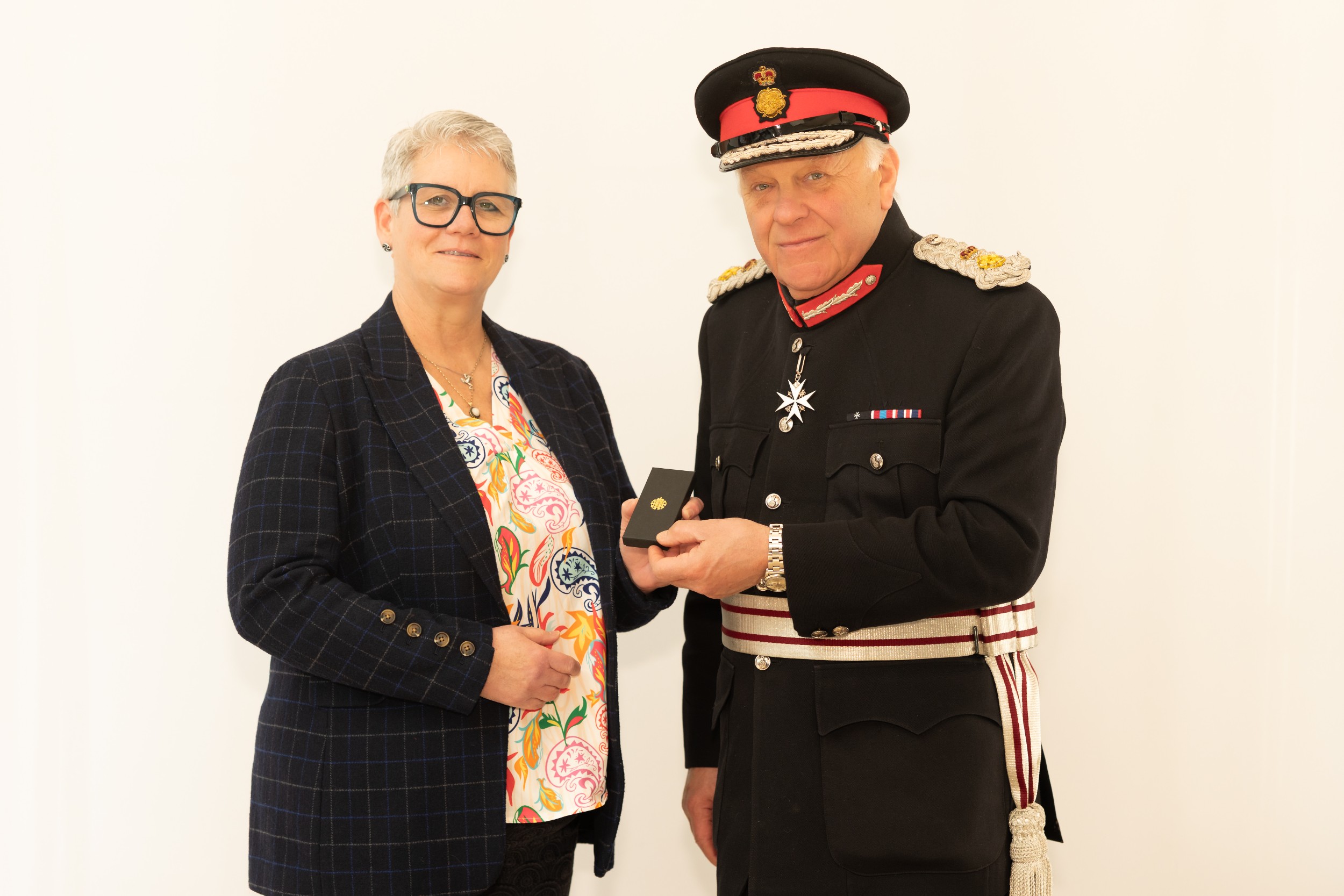 Gail Ladds in a patterned blouse and dark checked blazer stands beside Lord-Lieutenant Mr Toby Dennis wearing formal ceremonial uniform with medals and a peaked cap. He is presenting her with a presentation box as they stand against a plain light-coloured background