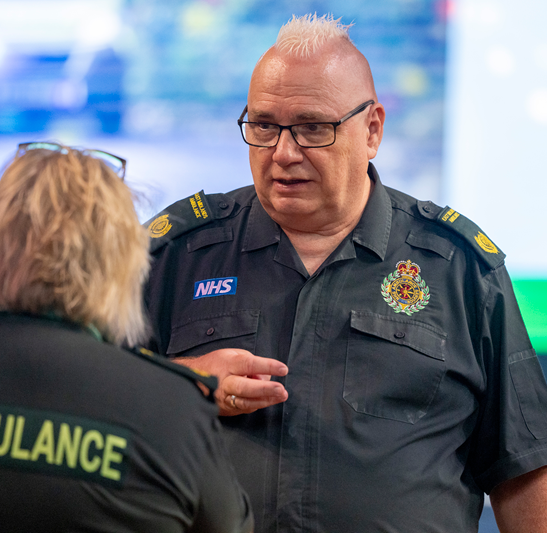 Simon Tomlinson, Assistant Director of Operations (EOC) at East Midlands Ambulance Service, speaks with a colleague during the award event while wearing an EMAS uniform.