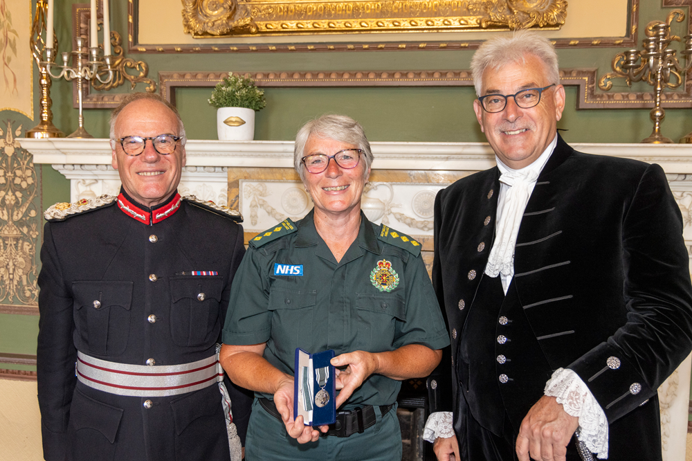 Helen Wootton stands in between the Lord-Lieutenant of Northamptonshire and High Sheriff of Northamptonshire. She is in green ambulance uniform and holds up her award.