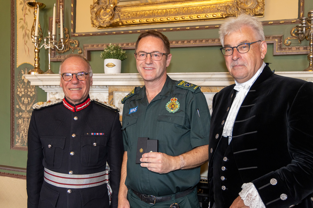 Aidan Ward stands in between the Lord-Lieutenant of Northamptonshire and High Sheriff of Northamptonshire. He is in green ambulance uniform and holds up his award.