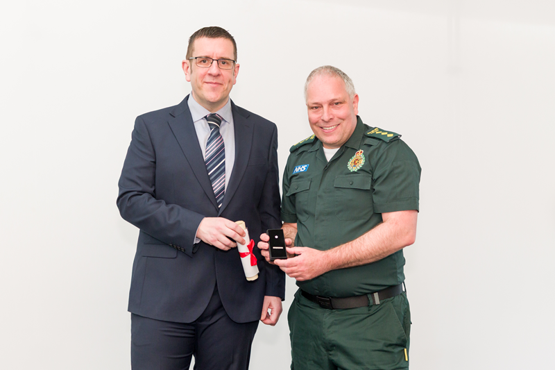 Mak Odam standing side by side with Ben Holdaway against a plain white background. Ben is wearing a dark business suit and tie. Mark is wearing green emergency service uniform and is holding his award, while Ben holds Mark's citation