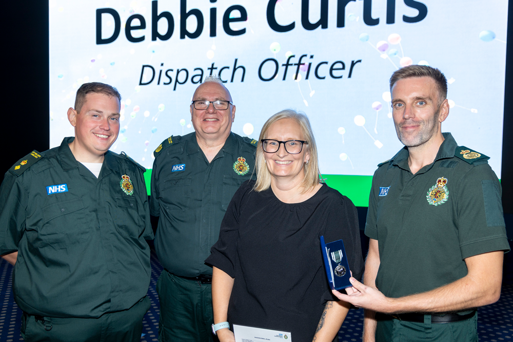 Four East Midlands Ambulance Service colleagues pose on stage at a long service awards event, with one colleague holding a medal presentation case. A large screen in the background displays the recipient's name and job title, "Debbie Curtis, Dispatch Officer".