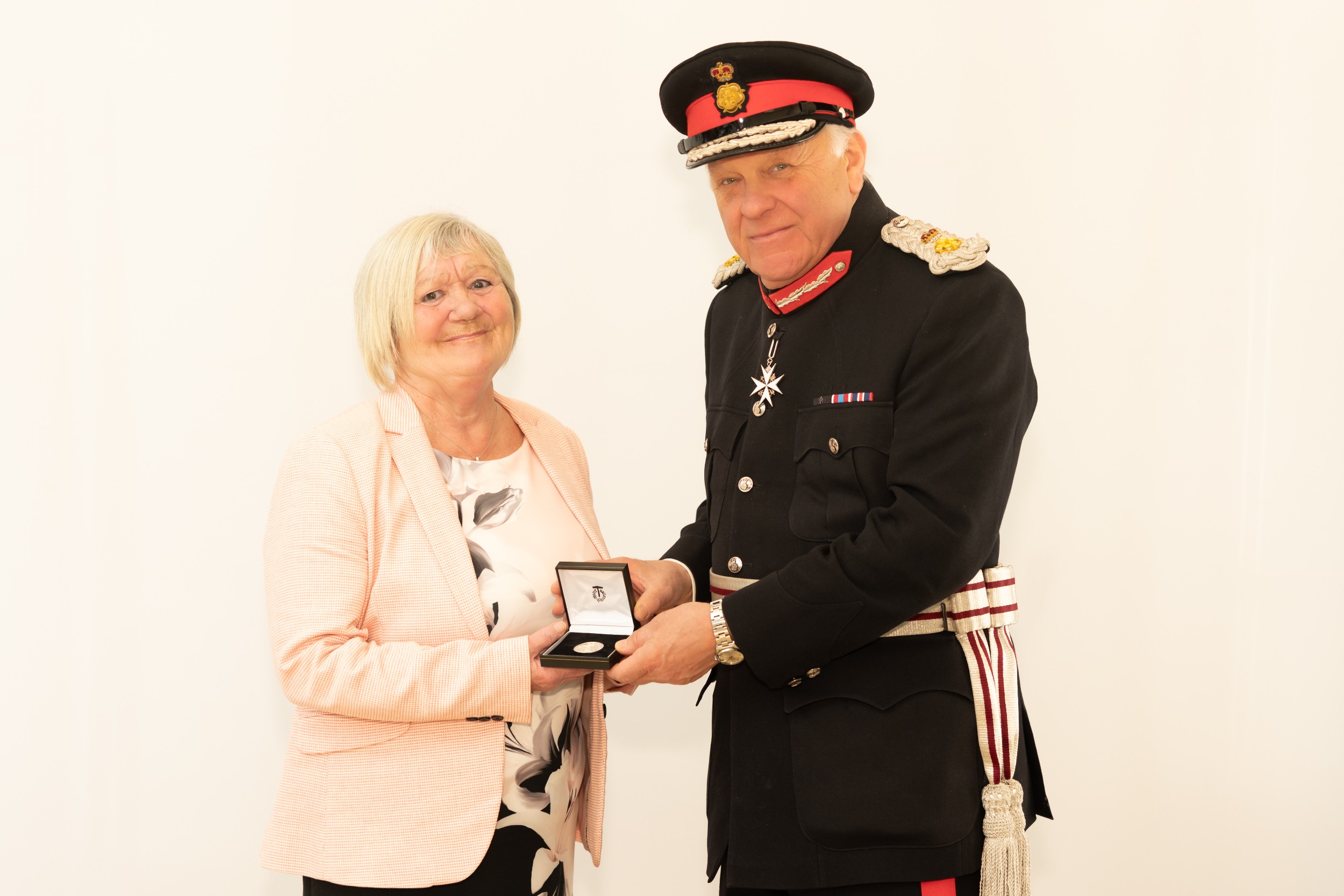 Pauline Dyer in a light patterned top and pale pink blazer stands beside Lord-Lieutenant Mr Toby Dennis dressed in a formal ceremonial uniform with medals and a peaked cap. He is presenting her with an open presentation box containing and award, and they stand against a plain light-coloured background.