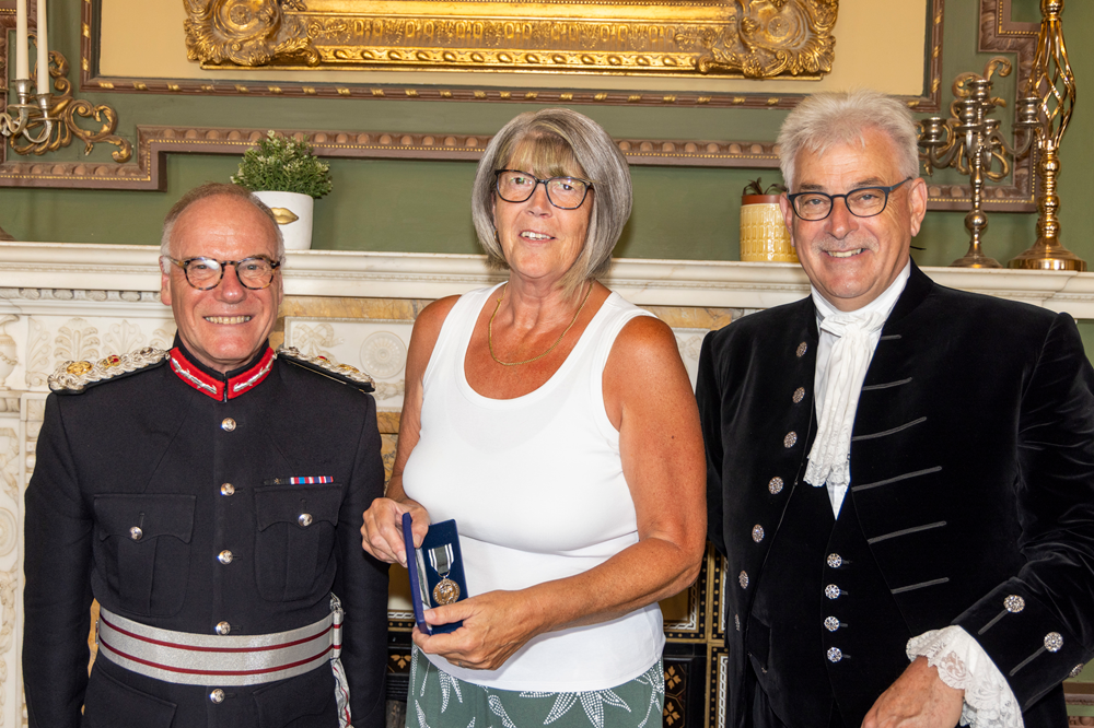 Paula Buchan stands in between the Lord-Lieutenant of Northamptonshire and High Sheriff of Northamptonshire. She is not wearing ambulance uniform and holds up her award.