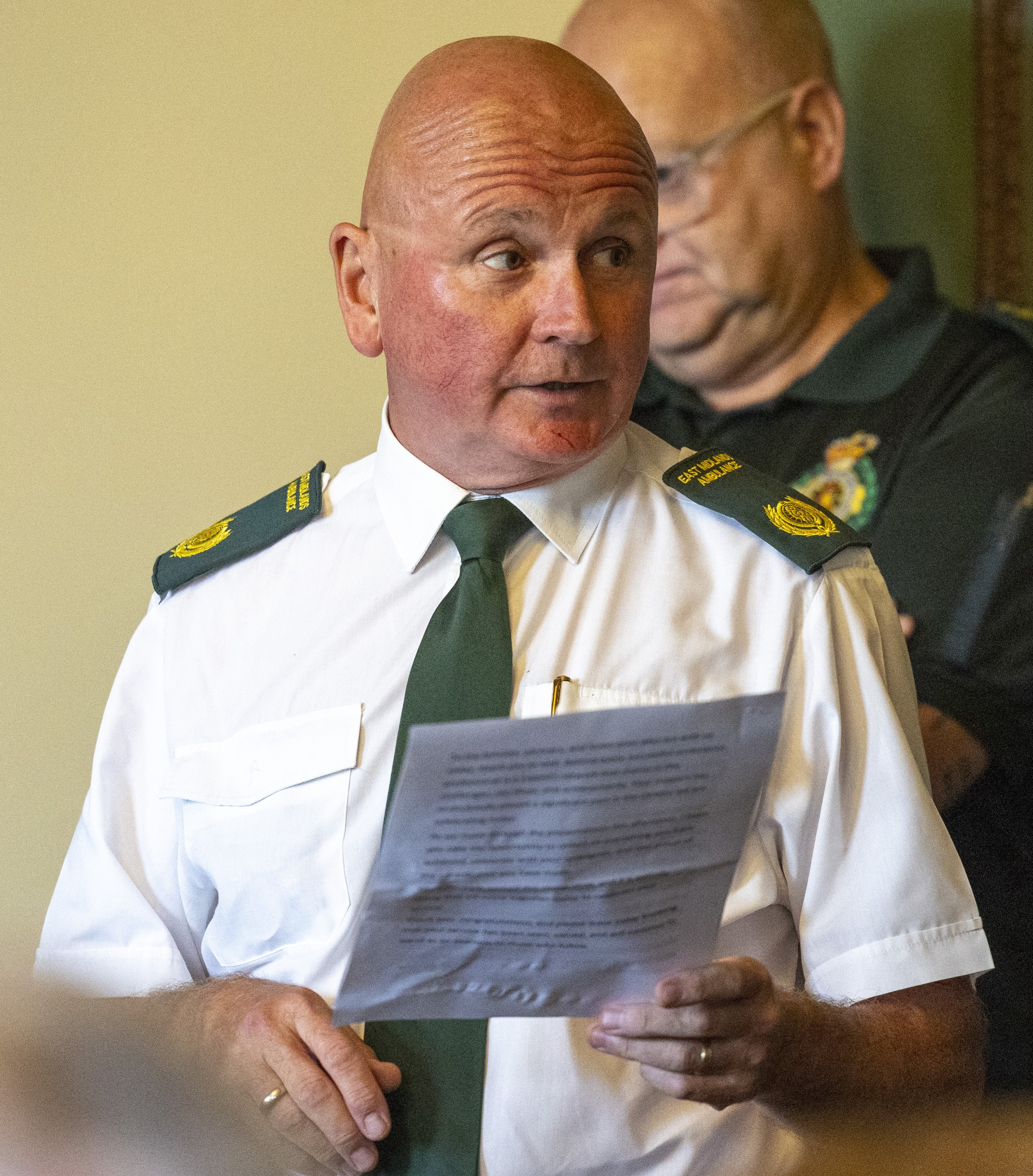 Mick Jones in a white shirt, with a green tie and epaulettes on his shoulders, addressing the attendees at the ceremony