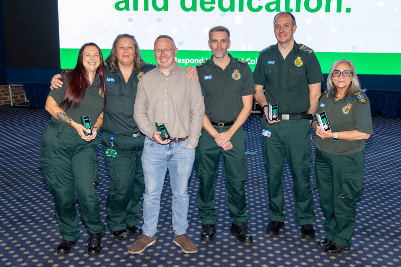 Six East Midlands Ambulance Service colleagues pose together on stage during a long service awards event, holding medal presentation cases. A large display screen is visible in the background as the group stands for a commemorative photograph.