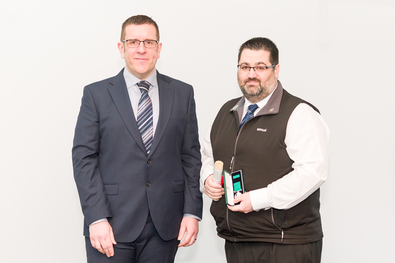 Ben Holdaway and Jake Marriott standing against a plain white background. Ben is wearing a dark business suit and tie. Jake is wearing a white shirt with a dark sleeveless zip up vest and is holding his award and citation.