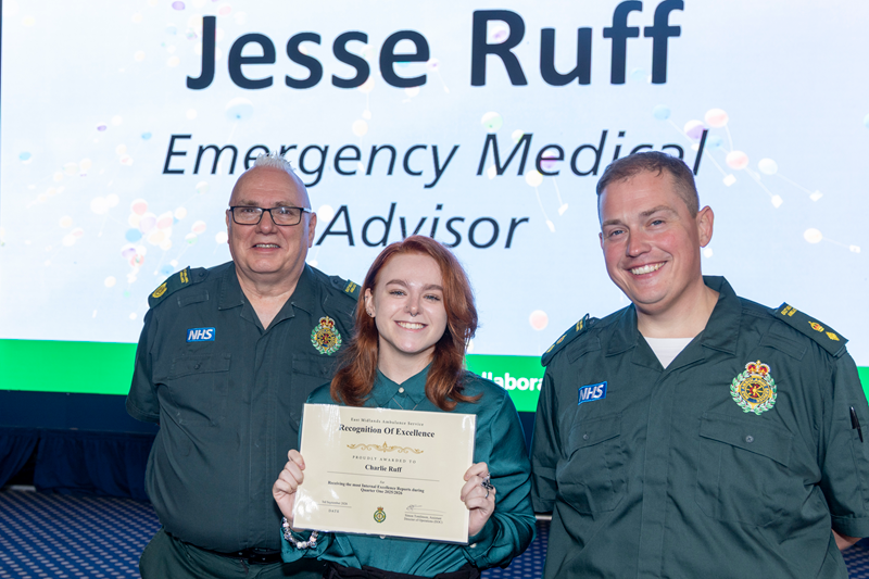 Three East Midlands Ambulance Service colleagues pose together on stage during an awards presentation. The central colleague holds a Recognition of Excellence certificate, while a large screen in the background displays the name "Jesse Ruff" and the job title "Emergency Medical Advisor".