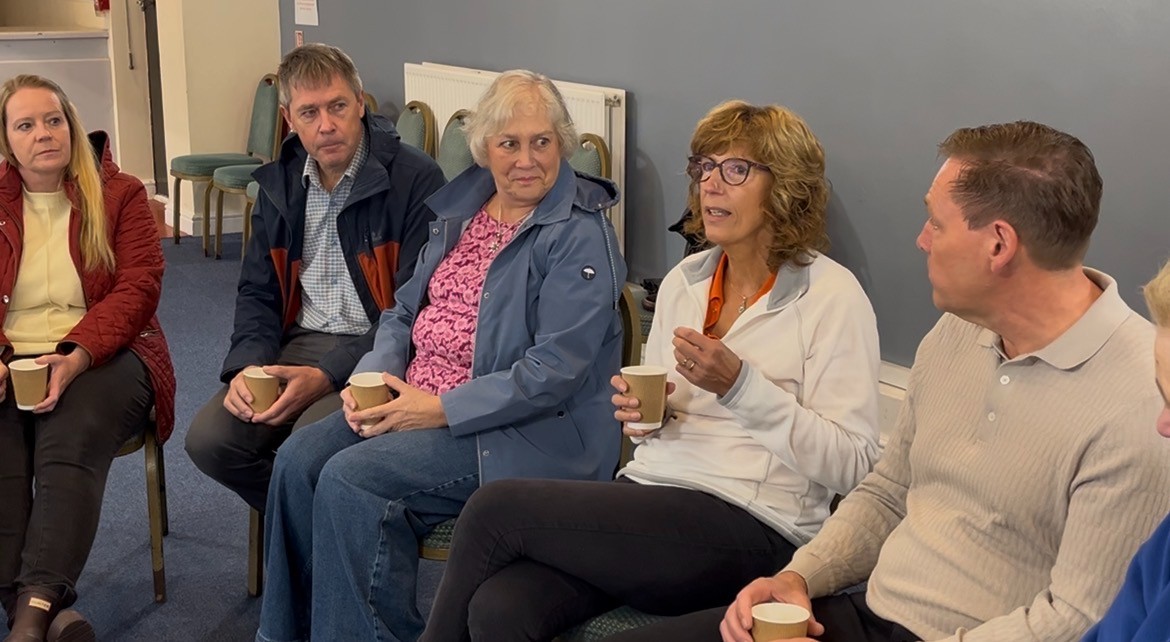 Anne has shoulder length brown hair and is wearing glasses. She holds a coffee while siting down and talking to her husband Peter, sitting to her right. To her left are Adele and James Lapworth and Stacey Wardle.