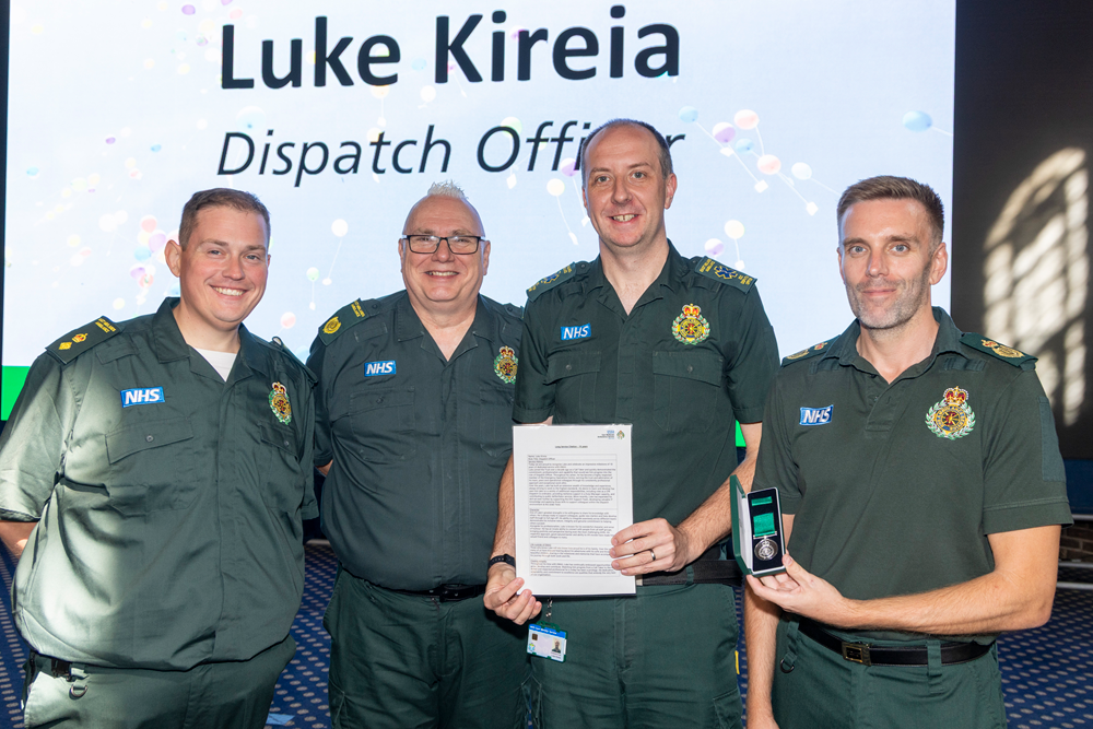 Four East Midlands Ambulance Service colleagues stand together on stage during a long service awards presentation. One colleague holds a framed certificate while another displays a medal in a presentation case. A large screen behind them shows the recipient's name and job title, "Luke Kireia, Dispatch Officer".