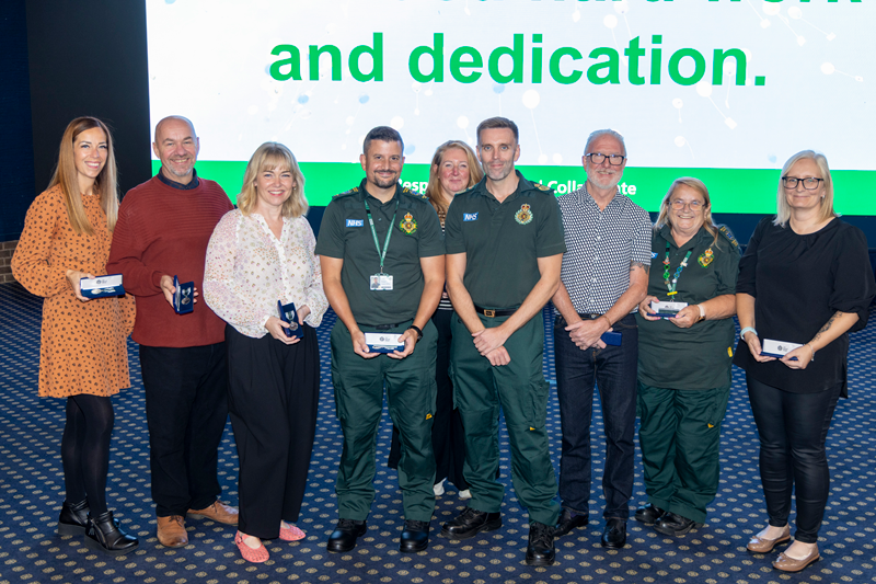 A group of East Midlands Ambulance Service colleagues and long service award recipients stand together on stage holding medal presentation cases.