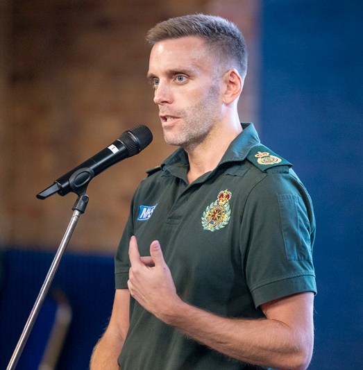 Ben Evans, Director of Service Delivery at East Midlands Ambulance Service, speaks into a microphone while addressing attendees at the event. He is wearing an EMAS uniform and standing on stage.
