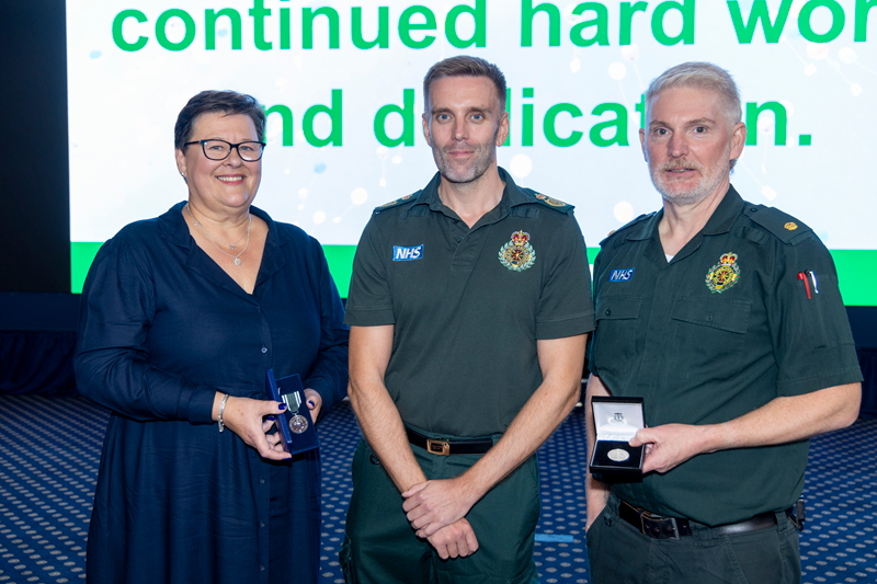 Three East Midlands Ambulance Service colleagues stand together on stage during a long service awards presentation. Two colleagues hold open medal presentation cases while a third stands between them. A large display screen can be seen in the background
