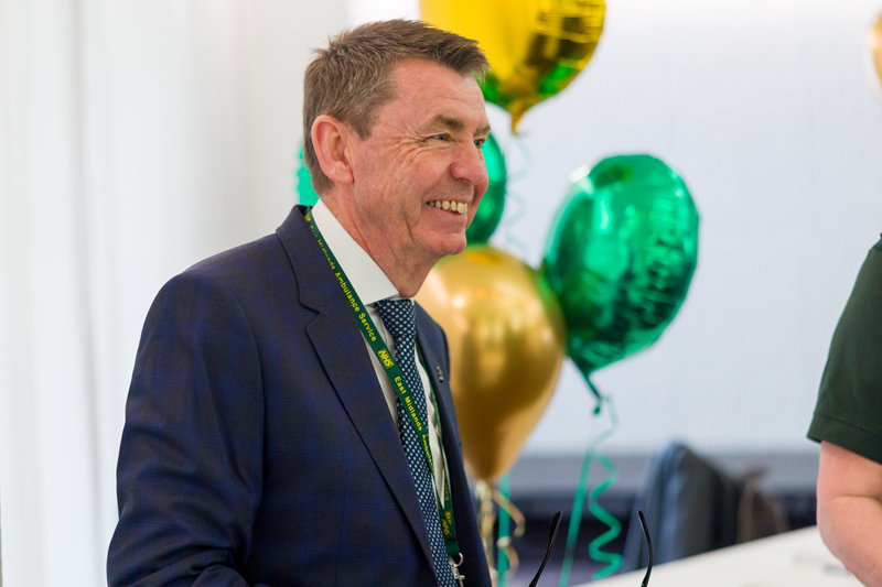Greg Cox wearing a dark suit, shirt, and tie stands indoors near a table. Green and gold balloons are visible in the background.