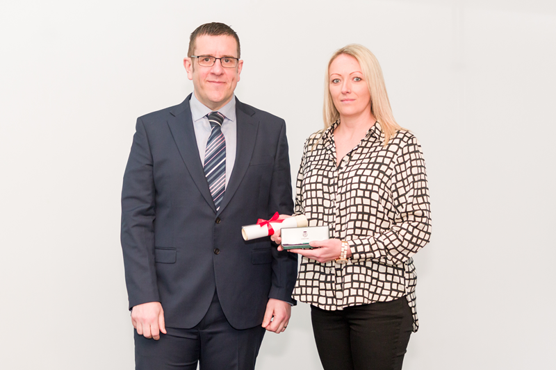 Kylie Raspin standing side by side with Ben Holdaway against a plain white background. Ben is wearing a dark business suit and tie. Kylie is wearing a patterned long-sleeve top and dark trousers, holding her award and citation. 