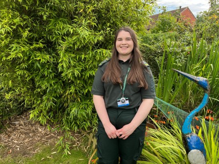 Chloe Irving stood smiling in a garden. There is an ornamental blue bird to her right. She is in her green uniform.