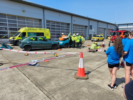 wide shot showing the simulation of a road traffic collision involving two cars. Emergency services staff attend to a patient in one of the cars while a crowd of student onlookers watch from afar.