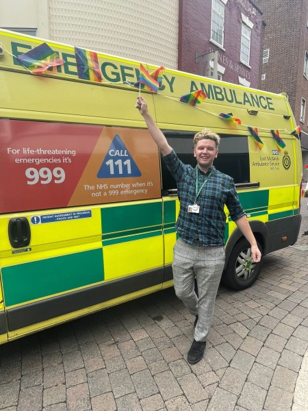 A man known as Anthony stands beside an EMAS ambulance waving a pride flag.
