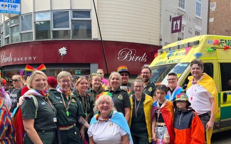 A diverse group of people from East Midlands Ambulance Service wearing rainbow costumes standing beside an ambulance.