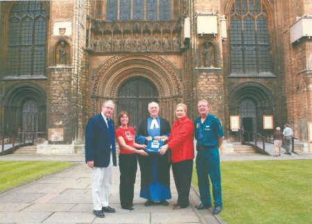 Nick Sentance in ambulance uniform standing with representatives from the British Heart Foundation and members of Lincoln Cathedral, with the cathedral in the background. They are holding up a defibrillator which was then installed in the Cathedral.