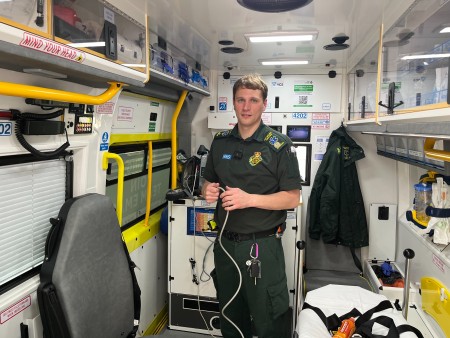 Dave Clarke standing in the back of the ambulance holding a cable. He has straight blonde hair and blue eyes. He is wearing his full ambulance uniform.