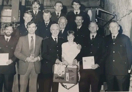 A younger Nick is pictured in a group photo with other colleagues in a black and white picture. At the front of the picture is a woman carrying the first defib received by EMAS in 1984.