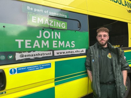 George Lane standing in front of our ambulance. He has brown curly hair and is wearing glasses. He is in his full green ambulance uniiform. 