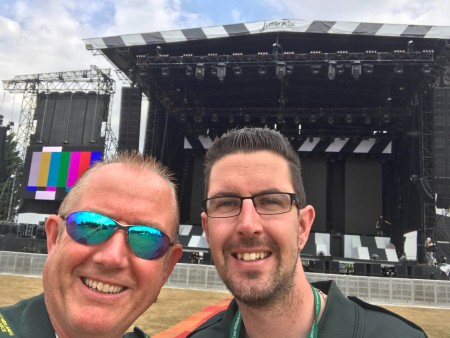 Brian and a male colleague posing for a selfie in front of an outdoor stage, where they are providing medical cover for an event..